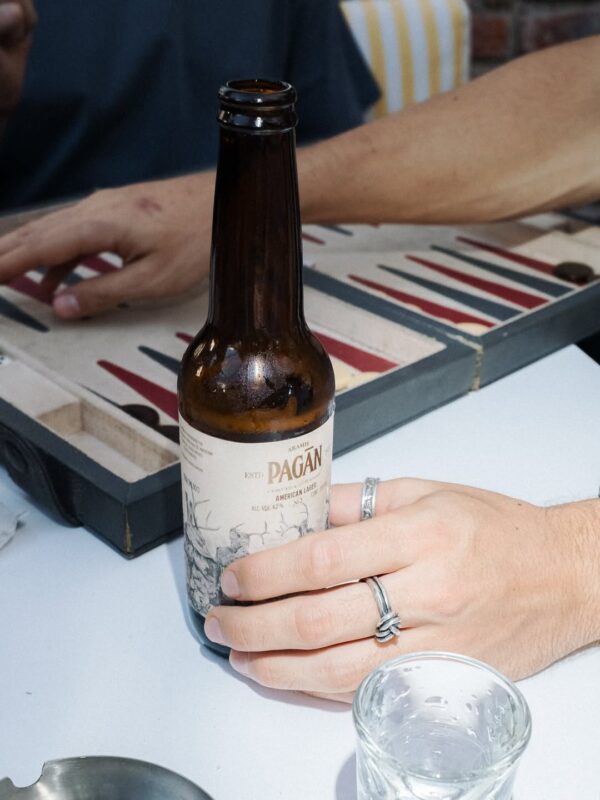 Persona sosteniendo una Cerveza Artesanal Pagān American Lager mientras juega backgammon, estilo lifestyle.